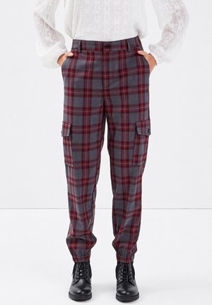 Grijs-rode geruite cargobroek met elastische manchetten, gedragen met zwarte veterlaarzen en een witte, gestructureerde blouse met lange mouwen.