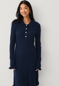Navyblaues geripptes Kleid mit kragenförmigem Ausschnitt, Knopfleiste und langen Ärmeln mit Rüschendetails. Weicher Stoff, figurbetontes Design.