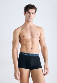 Zwarte katoenen boxer shorts met een blauwe elastische tailleband met het "Calvin Klein" logo. Het model heeft een neutrale houding, is shirtless en kijkt naar voren.