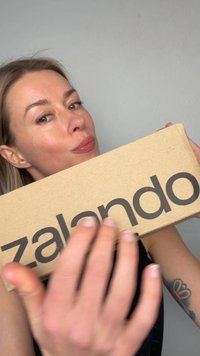 Vrouw met lichtbruin haar houdt een Zalando kartonnen doos dicht bij haar gezicht, gedeeltelijk verborgen door haar hand op de voorgrond.