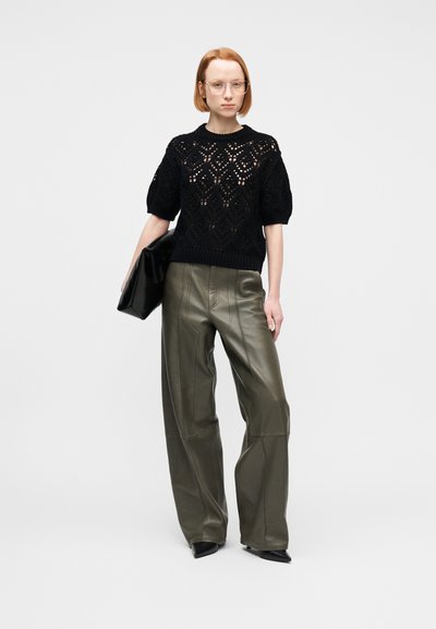 Γυναίκα με κοντά κόκκινα μαλλιά που φορά διαφανή γυαλιά, μαύρο κοντομάνικο πλεκτό τοπ, χακί φαρδιά δερμάτινα παντελόνια, μαύρα μυτερά παπούτσια, κρατώντας μαύρο clutch.