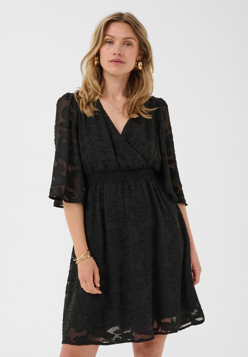 Femme portant une robe noire à motifs longueur genou avec des manches transparentes, des boucles d’oreilles créoles dorées, un collier et des bracelets sur un fond uni.