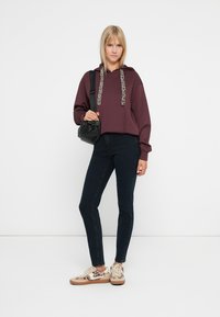 Ciemnofioletowa bluza z kapturem, ze sznurkami w print w cętki, w połączeniu z ciemnymi dżinsami i beżowymi sneakersami z wzorem w cętki. Czarna torba na ramię.