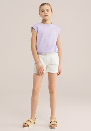 Jeune fille debout portant un haut sans manches violet clair avec un détail de palmier, un short blanc, des sandales beige et un bracelet violet.