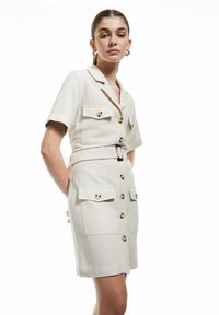 Beige jurk met korte mouwen en een ceintuur in de taille, twee borstzakken en gouden knoopdetails. Gemaakt van een lichte stof.