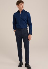 Camisa de vestir azul marino con cuello y botones, combinada con pantalones ajustados de un azul oscuro texturizado y zapatos de cuero negros.
