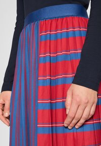 Jupe plissée présentant un schéma de couleurs rouge et bleu avec de fines rayures blanches horizontales. La ceinture est en tissu bleu uni.
