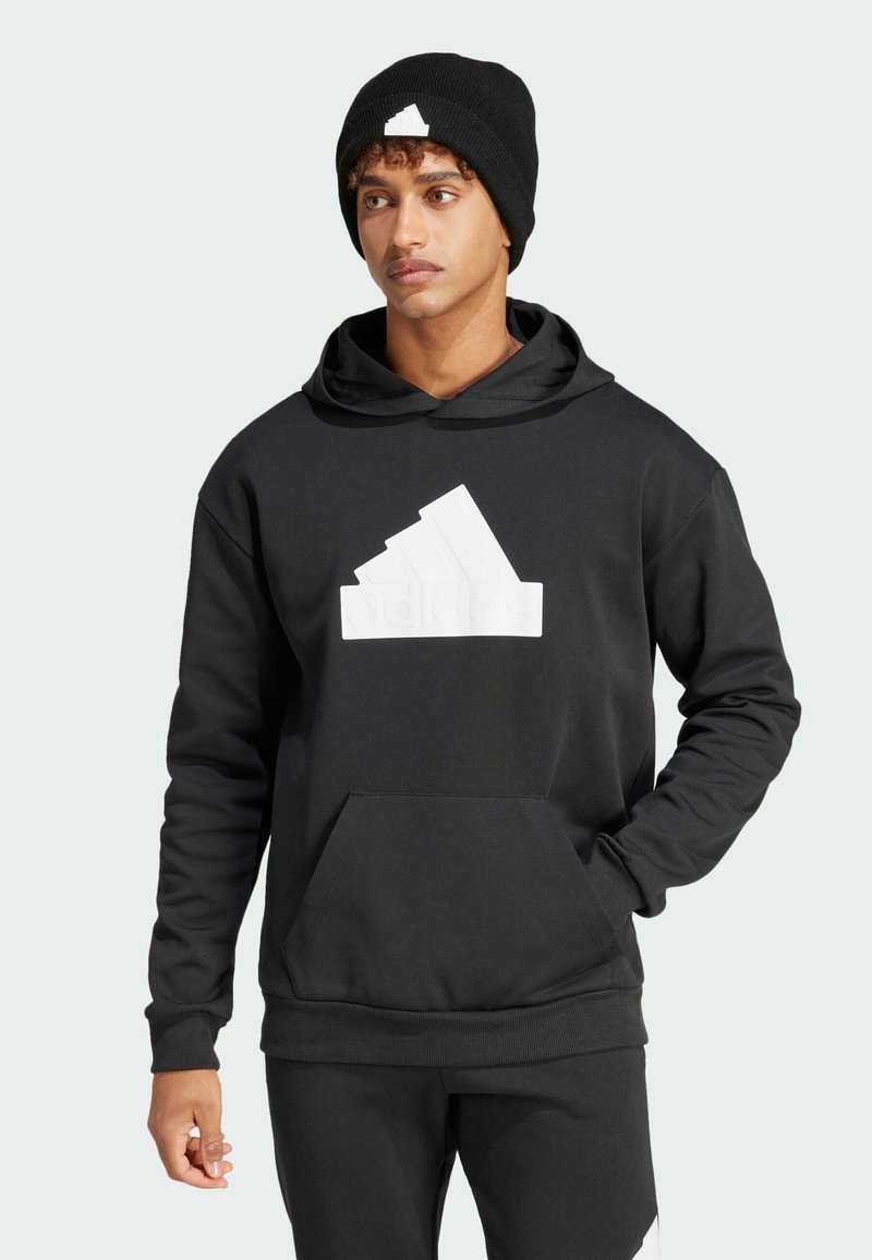 Sudadera negra con un gran logo blanco de Adidas, hecha de tela suave, con un bolsillo tipo canguro y puños acanalados. Llevada con un gorro negro.