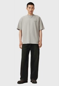 Mand iført en løs grå T-shirt, sorte bred benbukser, mørke sko og et tyndt sølv kædehalssmykke, stående foran en neutral baggrund.