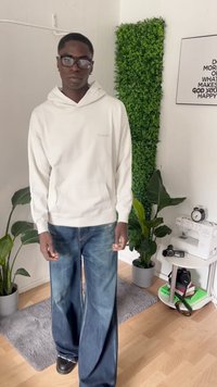 Sudadera blanca con capucha y bolsillo tipo canguro, con un logo gris, combinada con jeans de pierna ancha azules, en un entorno con plantas de interior y una máquina de coser.