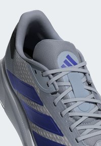 Gray athletic shoe with a textured surface, featuring purple accents, a mesh tongue, and gray laces. Includes a circular logo on the side.