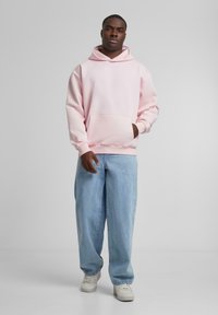 Lys rosa hettegenser med frontlomme og snøring, kombinert med løstsittende lyseblå jeans og hvite joggesko.