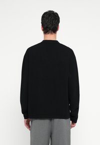 Μαύρο πλεκτό μπουφάν με υφή σχεδίου, με γιακά και μακριά μανίκια, συνδυάζεται με ανοιχτό γκρι παντελόνι. Δείχνεται από πίσω.