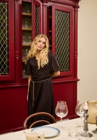Femme blonde en robe noire debout devant une armoire vintage rouge avec de la verrerie, à côté d'une table à manger dressée avec des assiettes et des verres.