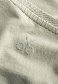 Lichtgrijze stof heeft een geborduurd scharenlogo in een iets donkerder grijs, wat een zachte textuur en eenvoudig ontwerp toont.