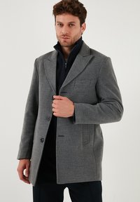 Cappotto in misto lana grigio con rever, chiusura a bottone, tasca frontale e maniche lunghe, indossato sopra un giubbotto interno in maglia blu navy.