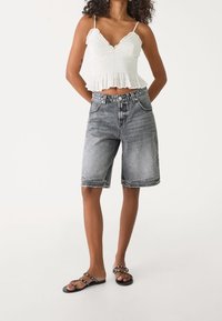 Witte, cropped, geplooide top met ruches aan de zoom; grijze denim shorts met vervaagde look; zwarte platte sandalen met kralenaccenten.