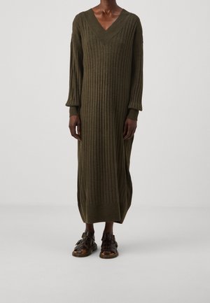 Długa, oliwkowozielona sukienka dzianinowa o prążkowanej fakturze, z głębokim dekoltem w kształcie litery V i długimi rękawami, uzupełniona paskowymi brązowymi sandałami.