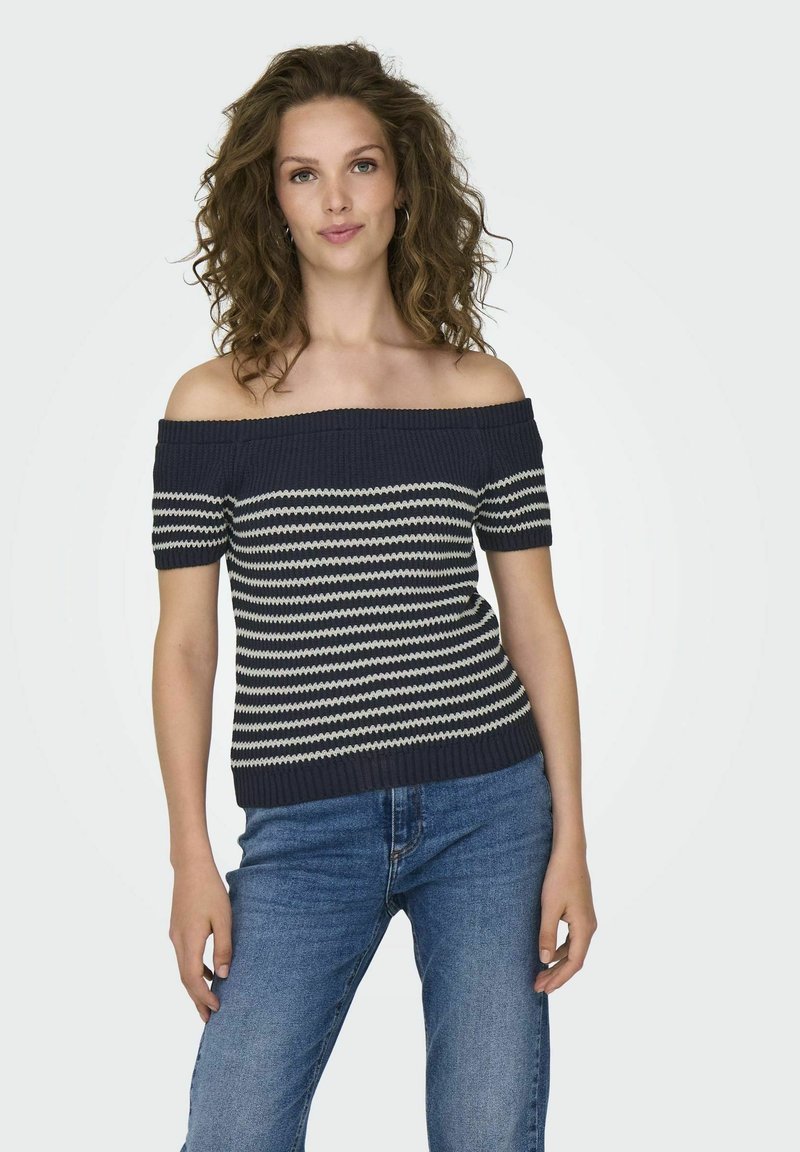 Top de punto off-the-shoulder con rayas en zigzag azul marino y crema, mangas cortas y textura acanalada. Combinado con jeans de mezclilla.