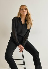 Blusa nera trasparente con colletto e chiusura a bottone, abbinata a pantaloni neri aderenti. La blusa ha maniche lunghe e un dettaglio con tasca.
