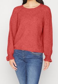 Roestkleurige gebreide trui met een geribbelde halslijn en losse, uitlopende mouwen. Bevat subtiele details en een licht verkorte lengte.