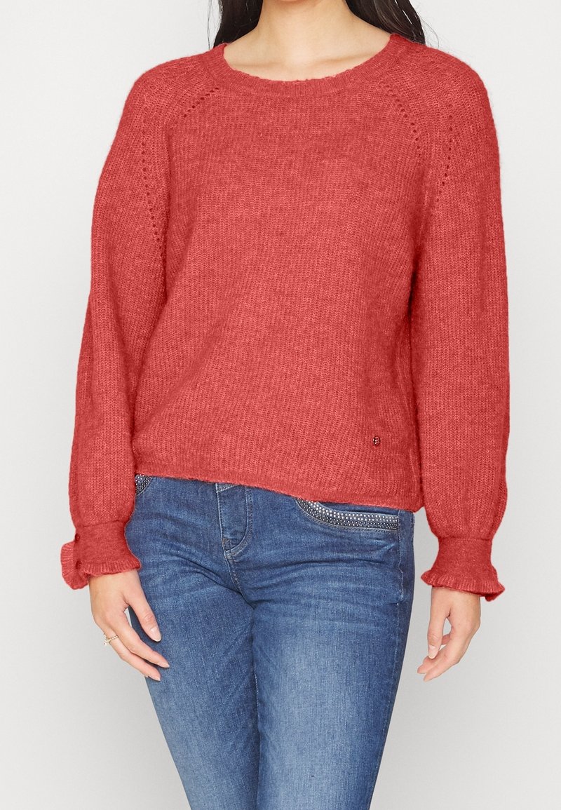 Roestkleurige gebreide trui met een geribbelde halslijn en losse, uitlopende mouwen. Bevat subtiele details en een licht verkorte lengte.
