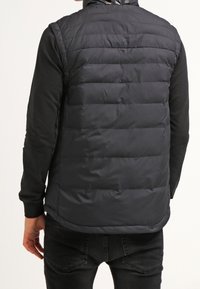 Vadderad svart väst med hög krage och horisontella sömmar, med en slät tygtextur. Bärs över en långärmad skjorta, bakvy visas.