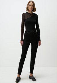 Body nero con maniche lunghe in rete e scollo asimmetrico, abbinato a jeans neri a vita alta. La modella indossa mocassini neri.