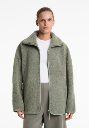 Jachetă din fleece de culoare verde deschis, cu guler înalt, închidere cu fermoar și croială supradimensionată. Textură netedă cu detalii de contrast în verde deschis.