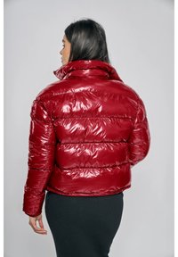 Röd vadderad jacka med glansig yta, utrustad med hög krage och horisontell vaddering. På baksidan finns en logotypetikett nära nacken.
