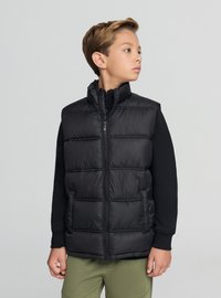 Czarna pikowana kamizelka z wysokim kołnierzem, zapinana na zamek błyskawiczny, z bocznymi kieszeniami. Noszona na czarnej bluzce z długim rękawem, zestawiona z zielonymi sportowymi spodniami.