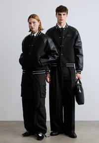 Jachete varsity negre cu mâneci din piele și dungi albe, asortate cu pantaloni cargo negri și o geantă neagră elegantă. Textură fină și design minimal.