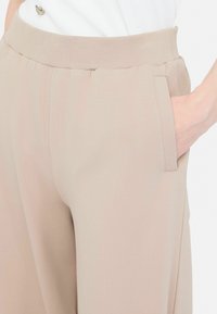 Pantaloni bej largi cu un brâu moale din tricot și buzunare laterale. Țesătura pare netedă și ușoară, reflectând o textură minimă.