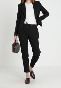 Zwarte, op maat gemaakte blazer over een witte top, gecombineerd met zwarte taps toelopende broeken. Accessoires zijn een ronde, gestructureerde handtas en bordeauxkleurige instappers.