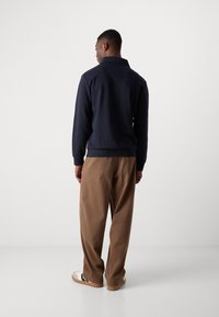 Sudadera navy con cuello, mangas largas, puños y dobladillo acanalados, combinada con pantalones marrones de corte holgado y zapatillas casuales.