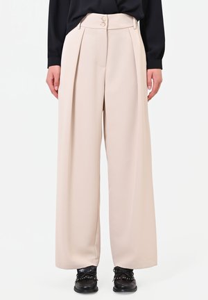 Pantaloni beige a vita alta e gamba larga con due bottoni, indossati con un top nero a maniche lunghe e scarpe nere con lacci.
