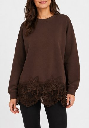 Femme portant un haut à manches longues marron foncé avec une bordure en dentelle florale à l'ourlet et un pantalon noir sur un fond uni.