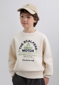 Krémově zbarvená mikina s výraznou černou a zelenou grafikou a textem, kulatý výstřih a žebrované manžety. Kombinována s béžovou čepicí.