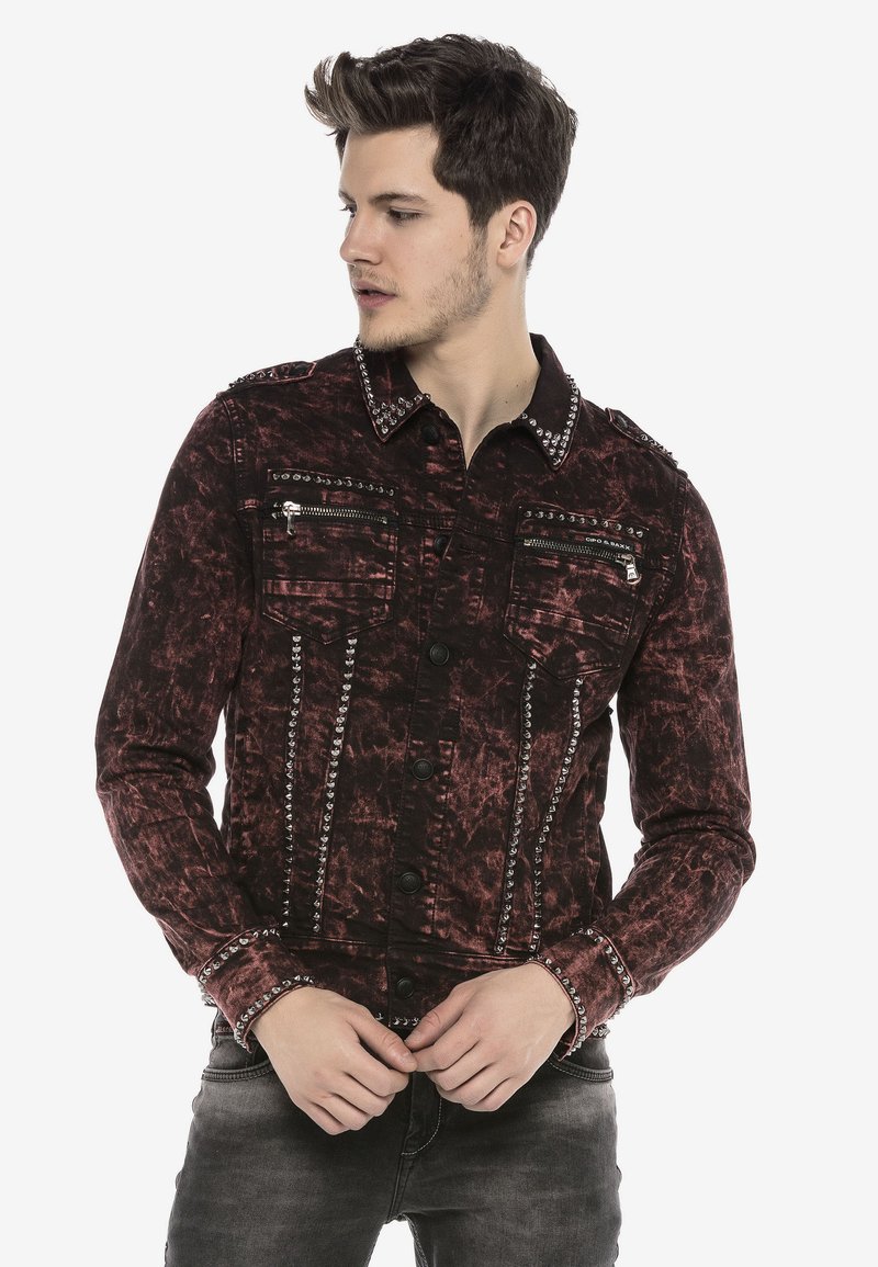 Rote und schwarze Jeansjacke mit einer verblassten, abgenutzten Textur. Verfügt über Reißverschlusstaschen auf der Brust, metallische Nieten entlang der Schultern und einen Knopfverschluss.