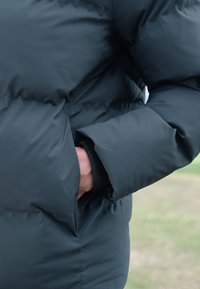 Person's hand inside the pocket of a black puffer jacket sleeve outdoors with a blurred grassy background.
