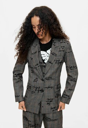 Vrouw met lang krullend haar die een grijs geruit pak draagt met zwarte handgeschreven tekst en een zwart-wit patroon trui.
