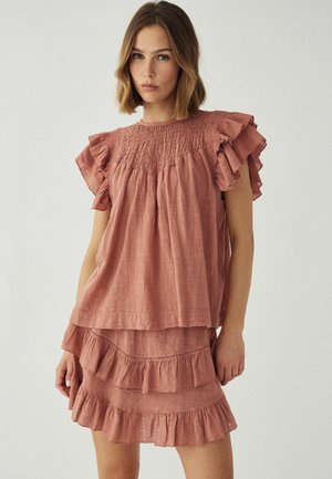 Kobieta ubrana w różową bluzkę z falbankami na krótki rękaw i pasującą plisowaną mini spódniczkę z falbankami, stojąca na jasnym, jednolitym tle.