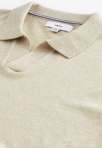 Beige polo skjorte med kort krave, prydet med en blå og hvid stribet detalje ved halsen, lavet af et blødt, struktureret stof. Mærketikette synlig.