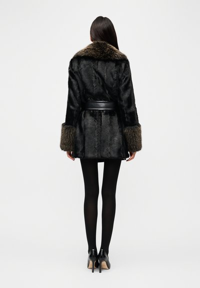Παλτό από συνθετική γούνα σε μαύρο με καφέ γούνινο γιακά, ζώνη στη μέση και υφασμένα μανίκια. Φορεμένο πάνω από μαύρα καλσόν και τακούνια.