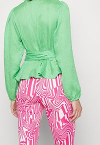 Blusa verde con texture, maniche a sbuffo e vita a peplum, abbinata a pantaloni rosa e bianchi con motivi a linee ondulate e forme geometriche.
