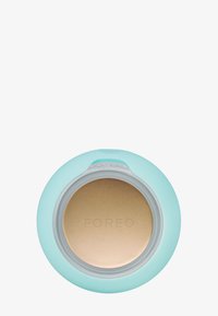 Rund kompakt med en ljusblå yta. Locket har en genomskinlig sektion. Inuti finns en bronzfärgad produkt med varumärket "FOREO" präglat.