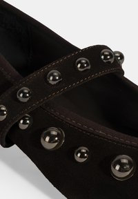 Schwarze Wildlederschuhe mit runder Form, ausgestattet mit zwei strukturierten schwarzen Riemen, die mit metallischen Nieten verziert sind. Weiche Innenauskleidung sichtbar.
