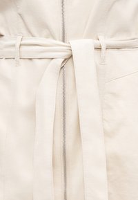 Εκρού ένδυμα με φερμουάρ στο μπροστινό μέρος και ζώνη από το ίδιο ύφασμα που δένεται γύρω από τη μέση μέσα από θηλιές για ζώνη.