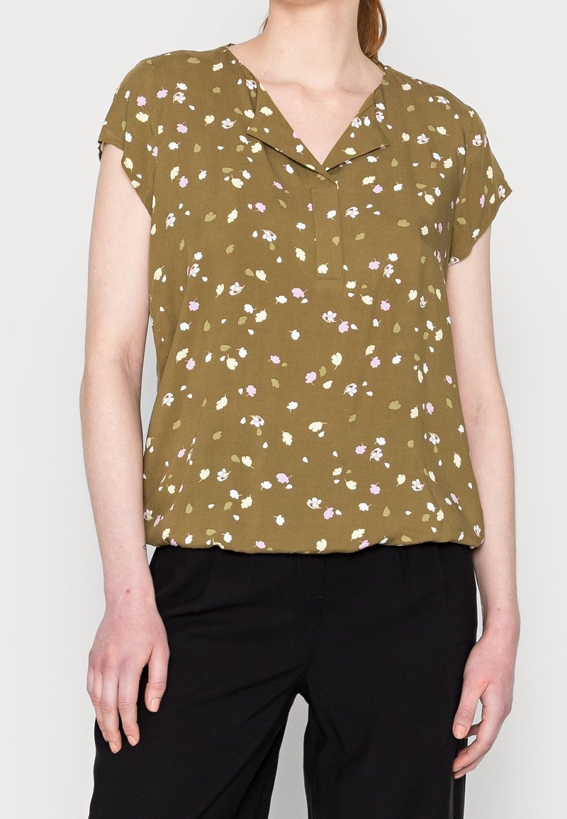 Personne portant un chemisier vert olive à manches courtes avec un petit imprimé floral blanc et rose, associé à un pantalon noir.