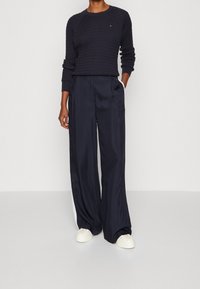 Marineblauer Strickpullover mit strukturiertem Muster, kombiniert mit marineblauen Satin-Hosen in weiter Beinform mit seitlichen Taschen und weißen Streifen. Weiße Sneakers.